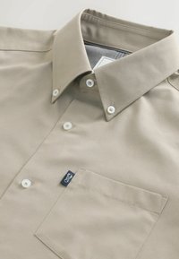 Beige gombos ing sima anyagból, hegyes gallérral, fehér gombokkal és egy zsebbel, amelyen egy kis logós címke található.