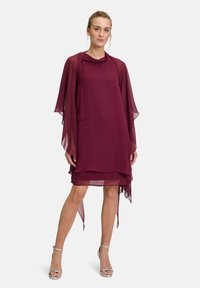 Burgunderfarbendes Chiffonkleid mit lockerer Passform, fließenden langen Ärmeln, geschichteter Optik und asymmetrischem Saum. Kombiniert mit silbernen Knöchelriemen.