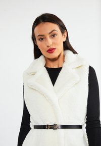 Witte fluffy vest met sjaalkraag, vastgemaakt met een zwarte riem met een gouden sluiting, gelaagd over een zwart langemouwen top.