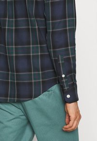 Camisa de cuadros azul marino y verde de manga larga con puños de botones blancos, sobre pantalones de color azul verdoso, con la mano derecha de una persona visible a su lado.