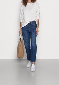 Beige mikina, tmavě modré džíny rovného střihu, bílé tenisky, drží béžovou tašku. Hladká tkanina, jednoduchý design a žádné viditelné vzory.