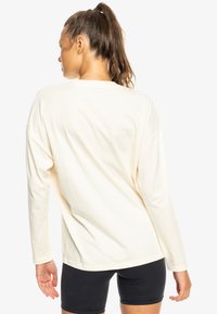 Camiseta de algodón crema de mangas largas con un corte relajado, escote redondo y costuras en los hombros caídos. Combinada con pantalones cortos negros.