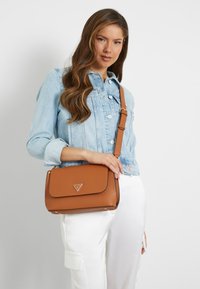 Borsa a tracolla in pelle marrone con flap e hardware dorato. Presenta un logo distintivo e una finitura testurizzata, da portare con una tracolla.