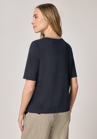 Kobieta o blond włosach, ubrana w granatową bluzkę z krótkim rękawem i beżowe spodnie o fakturze, zwrócona bokiem na jednolitym tle.