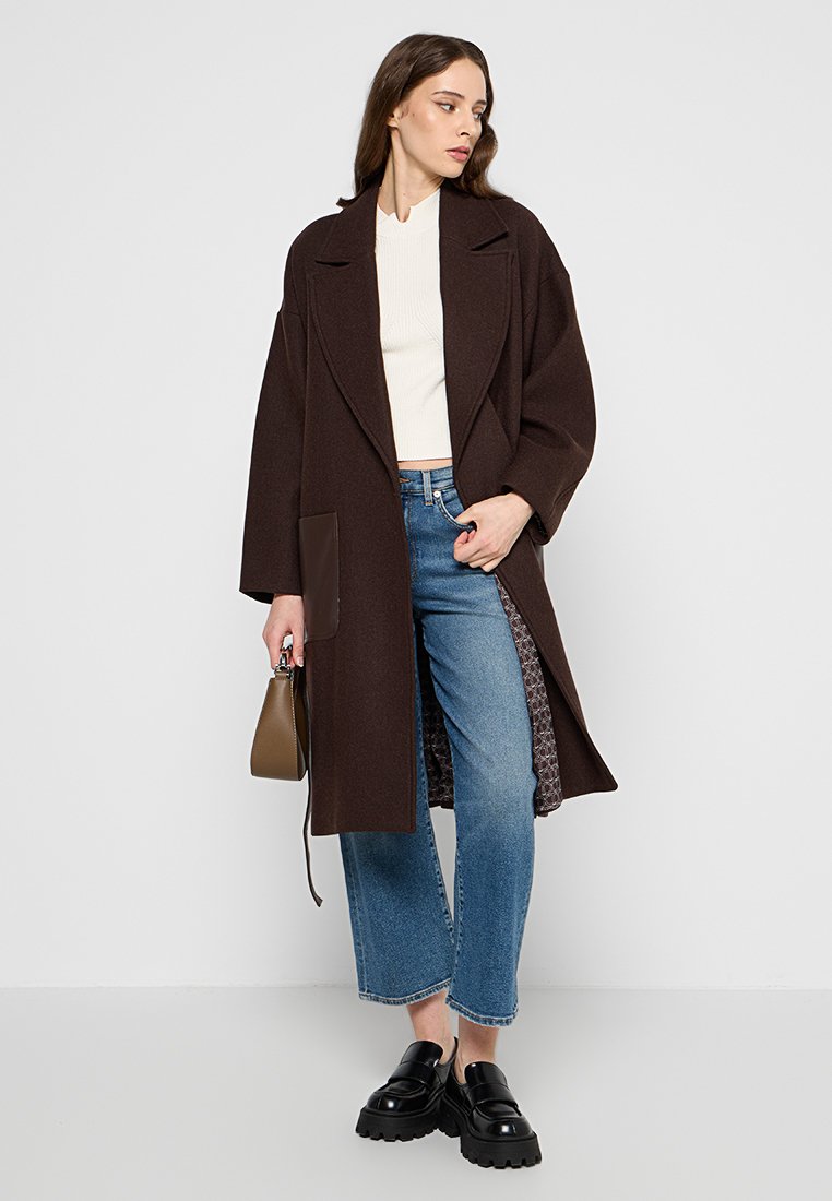 Brauner Wollmantel mit breiten Revers und einer einzelnen Tasche, über einem weißen gerippten Top und hellblauen Jeans getragen. Schwarze Plateauschuhe und Handtasche sind enthalten.