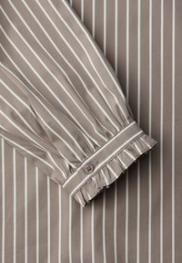 Chemise à manches longues rayée en taupe avec des rayures verticales blanches. Elle présente un poignet à volants froncés et une fermeture à un seul bouton. Tissu lisse.