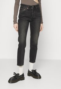 Jeans negros de cintura alta con corte recto, que cuentan con cinco bolsillos, costuras naranjas y un acabado desgastado, combinados con zapatos negros.