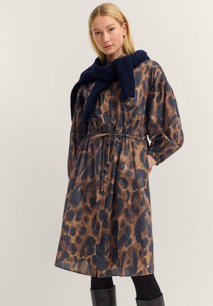 Frau in einem Midi-Kleid mit braunem und schwarzem Leopardenmuster, mit Kordelzug in der Taille, navyfarbigem Pullover über den Schultern, schwarzen Strumpfhosen und schwarzen Stiefeln.