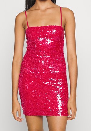 Woman wearing a fitted, pink sequin mini dress with thin shoulder straps, standing against a plain light background.