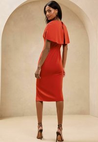 Punainen bodycon-mekko, jossa on viittamainen päällysosa, tiukka siluetti, polvimittainen helma ja avonainen selkäyksityiskohta. Yhdistetty remmi-korkokengillä.