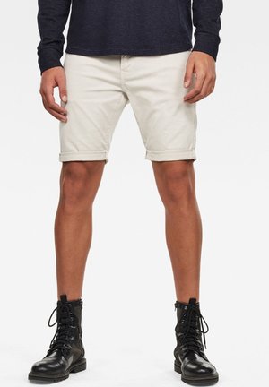 Homme portant un short beige retroussé, des bottes noires à lacets et une chemise sombre à manches longues, debout devant un fond uni.