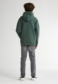 Zelená mikina s kapucí a volným střihem, spárovaná se šedými džíny a černobílými teniskami. Pohled zezadu, který ukazuje texturu a design.