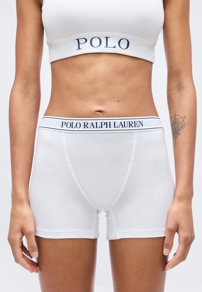 Γυναίκα με λευκό αθλητικό σουτιέν Polo Ralph Lauren και σορτς boxer, που δείχνει την κοιλιά και ένα τατουάζ στο αριστερό αντιβράχιο.