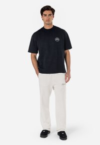 Mustal puuvillane T-särk lühikeste varrukatega ja ümmarguse kaelusega, millel on väike ringikujuline logo. Koos kreemika värvi dressipükste ja mustade kingadega.