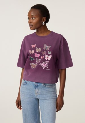 Vrouw draagt een paars T-shirt met roze en bruine vlinderprints en lichtblauwe spijkerbroek, kijkt naar links tegen een effen lichte achtergrond.