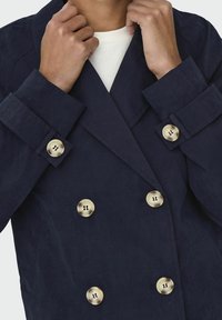 Giacca doppiopetto blu navy con grandi bottoni in ottone e tessuto testurizzato. Il colletto è largo con una vestibilità sagomata attorno alle spalle.