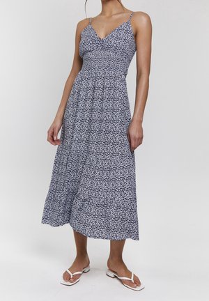 Femme portant une robe midi bleu marine à imprimé floral blanc, avec des bretelles fines et des sandales blanches à talons bas sur un fond uni.