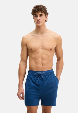 Hemdloser Mann mit lockigem Haar, der blaue Shorts mit Kordelzug und Marc O’Polo-Bund trägt, steht vor einem schlichten hellen Hintergrund.