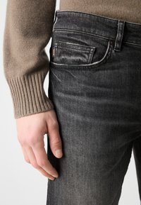 Jeans din denim gri, cu o croială slim, având un design clasic cu cinci buzunare. Materialul prezintă o textură ușoară, cu cusături vizibile și eticheta brandului.