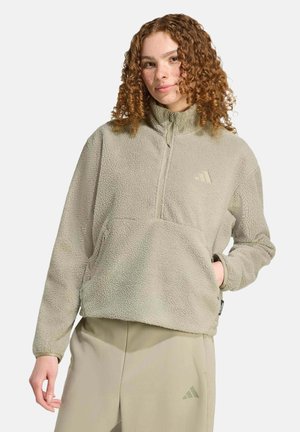 Giovane donna con capelli ricci che indossa una giacca in pile beige chiaro e pantaloni abbinati con logo, in piedi davanti a uno sfondo neutro.