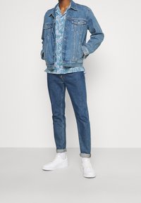 Chaqueta vaquera azul claro, camisa de manga corta con estampado floral, vaqueros oscuros estrechos y zapatillas blancas. Presenta puños remangados y bolsillos.