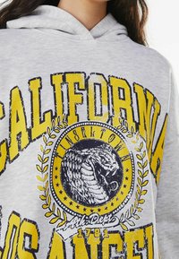 Světlé šedé mikina s kapucí se žlutým a černým grafickým potiskem. Design obsahuje text "CALIFORNIA" a kruhový emblém hada s vavřínovými akcenty.
