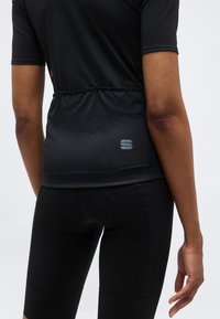 Άτομο που φορά μαύρη ποδηλατική φανέλα με κοντά μανίκια και πίσω τσέπες, και μαύρο επενδυμένο ποδηλατικό σορτς, στέκεται μπροστά σε απλό ανοιχτό φόντο.