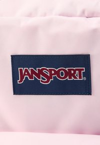 Gros plan sur un écusson rectangulaire JanSport bleu foncé avec texte blanc et rouge foncé sur un fond de tissu texturé rose clair.