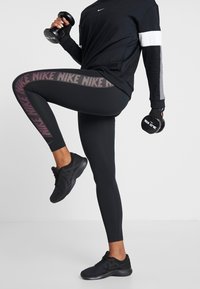 Czarny długi rękaw sportowy top z szarymi i białymi akcentami. Czarna legginsy z wzorzystą boczną lamówką. Model trzyma ciężarki, ubrany w czarne trampki.