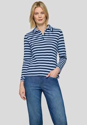 Blonde woman stands against gray background wearing blue and black striped long-sleeve polo shirt and blue jeans, one hand in pocket.