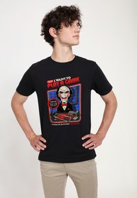 Camiseta de algodón negra con un gráfico de Jigsaw de "Saw". El diseño incluye acentos rojos y azules, texto y elementos de temática de horror.