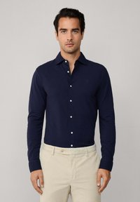 Man in een strakke marineblauwe lange mouwen overhemd met knopen, gestoken in beige broeken, staand tegen een effen lichte achtergrond.