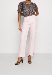 Blouse noire fleurie avec manches bouffantes, détail œillet ; jeans roses évasés avec fentes latérales ; sandales à talons noires. Design texturé et décontracté.