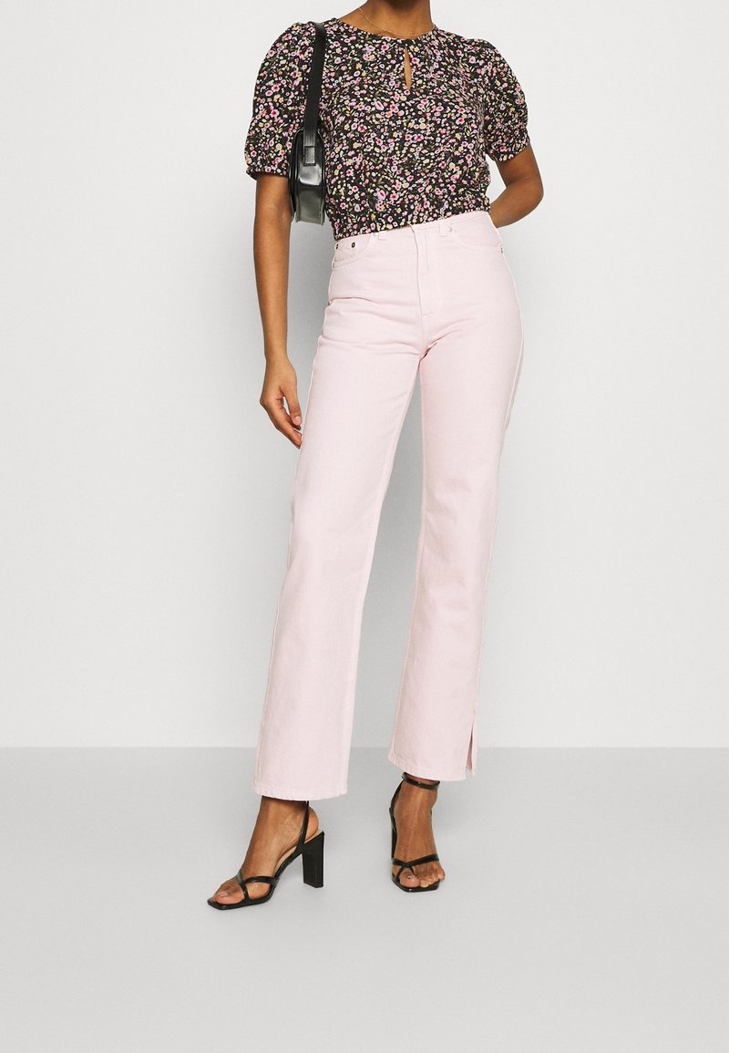 Blouse noire fleurie avec manches bouffantes, détail œillet ; jeans roses évasés avec fentes latérales ; sandales à talons noires. Design texturé et décontracté.