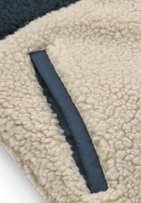 Detail van een zak van een beige en donkere fleecejas van stof, met een getextureerde wolachtige buitenkant en een gladde binnenvoering van de zak.