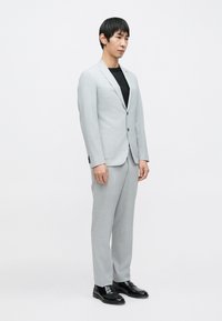 Világosszürke öltöny strukturált zakóval, hozzáillő nadrággal és fekete cipővel. A szövet finom mintázatú; a fazon szabott és modern.