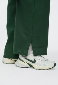 Biele a zelené tenisky Nike nosené s tmavozelenými širokými nohavicami, pričom je vidieť malý zárez na okraji nohavíc nad bielymi ponožkami.