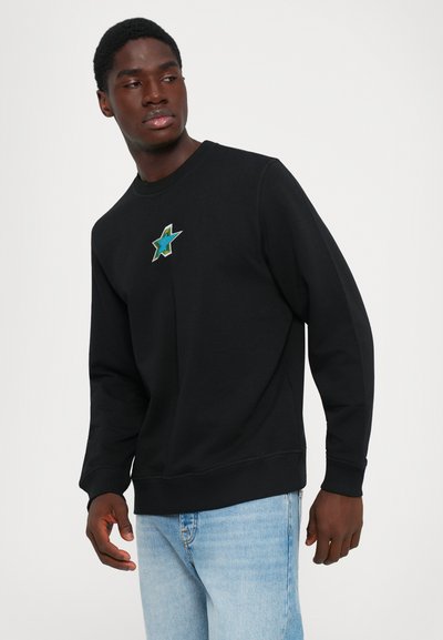 Man wearing black sweatshirt with blue star design on chest and light blue jeans, looking to his left against plain background.