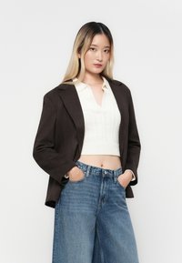 Brūns žakets no gludas auduma, valkāts pār krēma krāsas pamatā adītutopu un augsta jostasvietā ziliem džinsiem. Modelis pozē ar rokām kabatās.