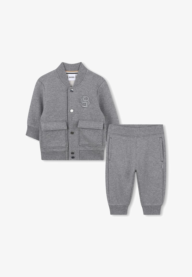 Giacca grigia per bambino con maniche lunghe e bottoni, dotata di due tasche frontali e pantaloni coordinati con polsini elasticizzati, con logo in rilievo sul petto della giacca.