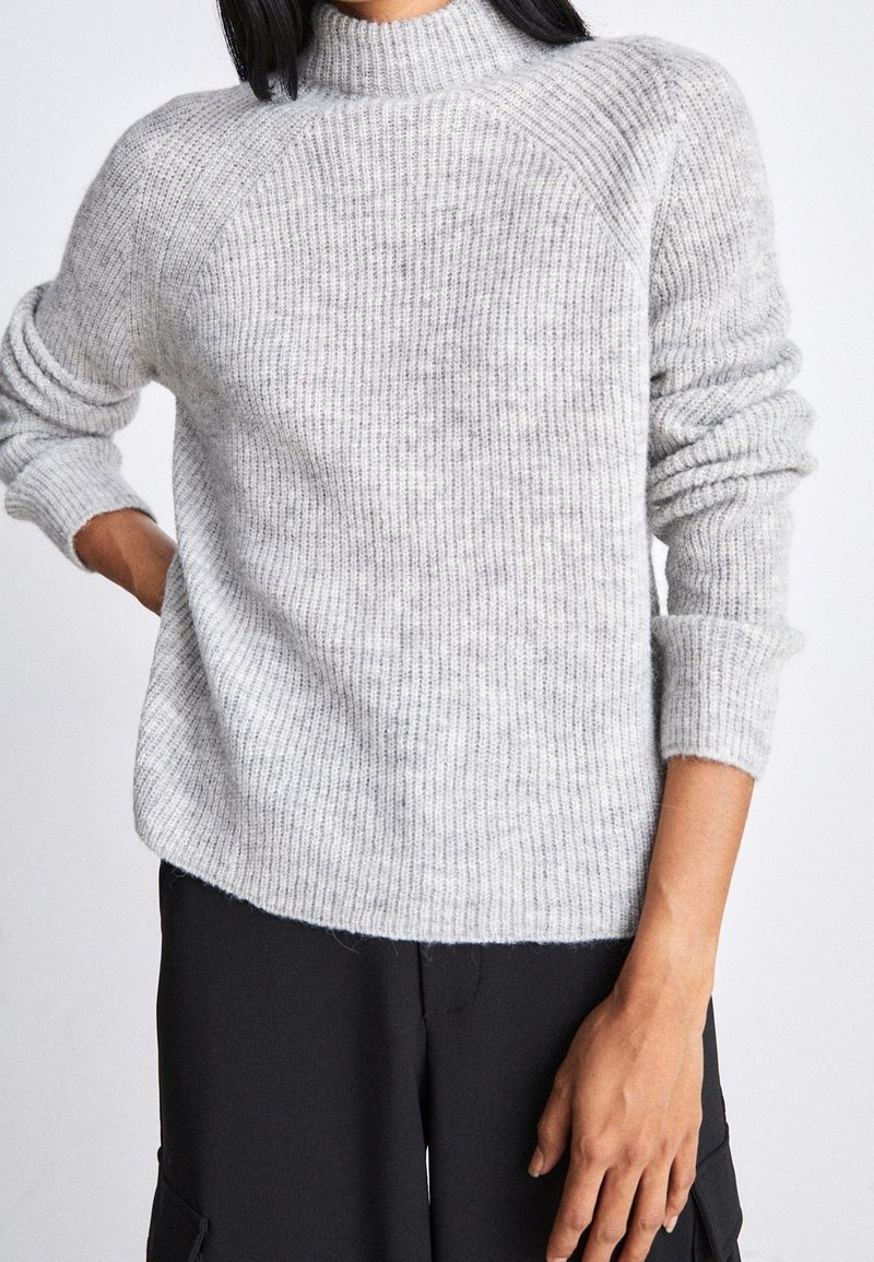 Personne portant un pull à col roulé côtelé gris clair à manches longues et un pantalon noir, debout avec une main sur la hanche.