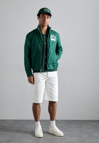 Grønn jakke med glidelås og logo, sammen med en mørk knappeskjorte, hvite shorts, hvite sneakers og en matchende grønn caps.