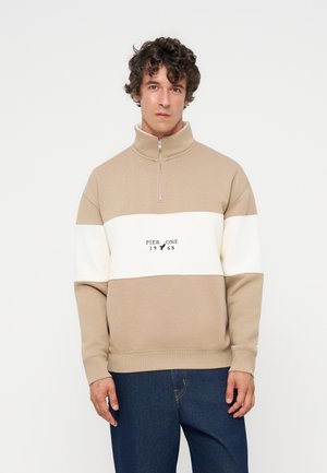 Homme aux cheveux bruns bouclés portant un sweat-shirt beige et blanc avec fermeture quart de zip avec le texte "Pier One 1968" et un jean bleu.