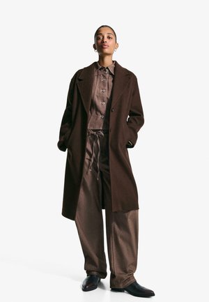 Femme debout avec les mains dans les poches, portant un manteau long marron, une chemise à boutons, un pantalon large et des chaussures noires sur fond blanc.