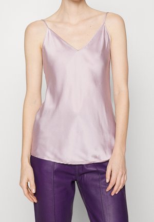 Nainen, joka pukeutunut vaaleanpunaiseen satiiniseen topiin ohuilla olkaimilla ja korkeavyötäröisiin violetteihin nahkahousuihin, seisoo yksiväristä taustaa vasten.