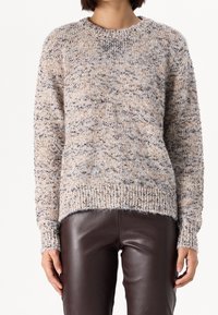 Strickpullover in Beige und Braun mit einem strukturierten, gesprenkelten Muster. Verfügt über einen Rundhalsausschnitt sowie gerippte Bündchen und Säume. Wird mit braunen Lederhosen getragen.