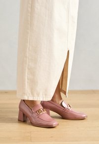 Rosa Lackleder-Pumps mit Absatz und goldener Kettenakzent, mit abgerundeter Zehenform und Blockabsatz, kombiniert mit einem cremefarbenen Rock.