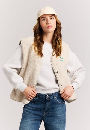 Junge Frau trägt beigen Fleeceweste, hellen Sweatshirt, blaue Jeans und beige Kappe und steht vor einem einfarbigen Hintergrund.
