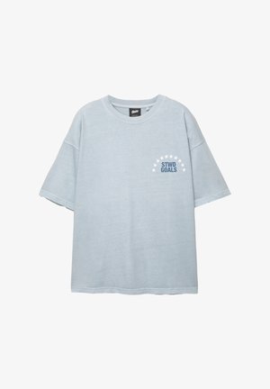 Vaaleansininen lyhythihainen T-paita, jossa on pyöreä pääntie. T-paidassa on "STWD GOALS" -teksti, joka sijaitsee vasemmassa rinnassa valkoisten tähtien puoliympyrän alla.