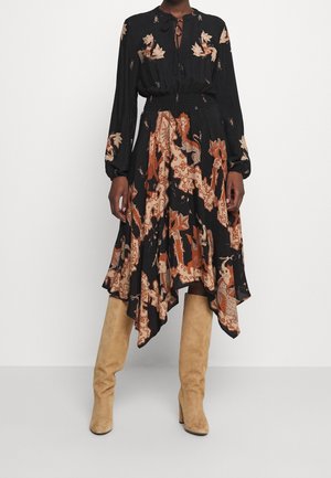 Frau trägt ein langärmeliges Kleid mit schwarz-braunem Muster und Tuchsaum sowie beige kniehohe Wildlederstiefel.